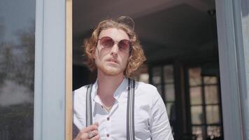 retrato do uma jovem masculino modelo dentro oculos de sol deixando cafeteria e caminhando em uma varanda. Ação. jovem homem dentro branco camisa perto a construção em uma verão ensolarado dia. video