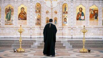 el iconostasio dentro un ortodoxo iglesia. . posterior ver de un sacerdote en pie en frente de el íconos con el caras de el santos, concepto de religión, interior detalles dentro un iglesia. video