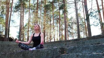 Woman practises meditation surrounded by pine forest. Concept. Young spiritual girl meditates outdoors sitting on stone stairs. video