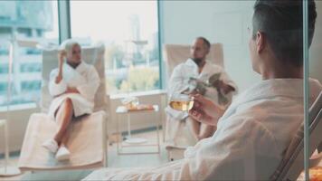 People relaxing at the spa salon. . Two men and a woman in white bathrobes having a rest with tea, newspaper inside a spacious room with panoramic windows. video