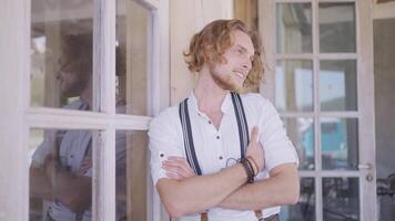 A man in white shirt with curly blond hair near countryside house. Action. Handsome young man standing in front of the windows of a beautiful house with glass dorrs and smiles. video