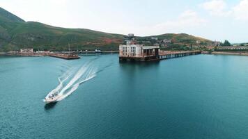 Aerial view of a motorboat moving away from a picturesque pier. Shot. Summer landscape with a coastal city and green mountains. video