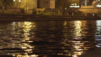 schließen oben von das Oberfläche von das Wasser mit das Beleuchtung von das Nacht Stadt. Lager Filmaufnahme. wellig breit rive und das dunkel Stadt Straße auf ein Sommer- Nacht mit Gehen Personen. video