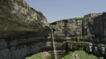 Narrow waterfall falls from high cliff. Action. Top view of water from hand falling into waterfall. River falls and becomes waterfall in mountains. Beautiful rocky cliff with narrow waterfall video