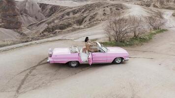 bello giovane caucasico sposa e sposo durante sessione fotografica nel tacchino, cappadocia. azione. aereo Visualizza di appena sposato coppia nel un' rosa Vintage ▾ cabriolet auto su il sfondo di roccia formazioni. video