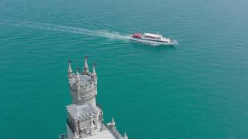 Castle on background of yacht in blue sea. Action. Top view of beautiful white castle near sea cliff with floating yacht. Yacht sails near white castle over bluff of blue sea. Swallow's nest in Crimea video