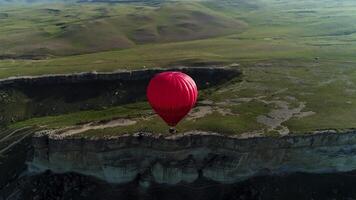 antenn panorama- se av en enda röd varm luft ballong flygande ovan fantastisk klippa täckt förbi grön äng. skott. lantlig sommar landskap med de ändlös grön fält och blå horisont. video