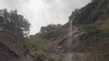 parte inferiore Visualizza di un' piccolo cascata e un' ripido scogliera su nuvoloso cielo sfondo. azione. bellissimo verde montagna pendenza e un' cascata. video