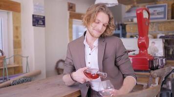 A handsome man with curly blond hair in beige suit pouring rose wine into a transparent glass. Action. Male sommelier pouring fruit wine at a bar. video