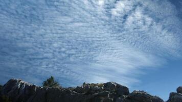 un' chiaro leggermente nuvoloso cielo con un' modello. creativo. contro il sfondo di il cielo è un' bellissimo grigio montagna con un' piccolo numero di alberi e grigio pietre sotto. video