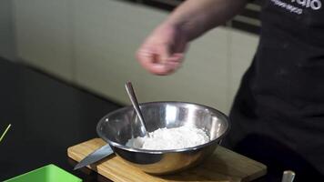 fechar acima do masculino mãos adicionando sal para dentro a farinha para amassar massa. arte. mistura ingredientes dentro uma metal tigela colocada em uma de madeira borda com uma colher. video