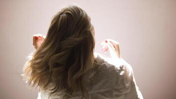 ein jung Frau zittern ihr blond lockig Haar isoliert auf ein Beige Mauer Hintergrund. Kunst. Rückseite Aussicht von ein jung blond Frau im ein Weiß Hemd posieren und berühren ihr Haar. video