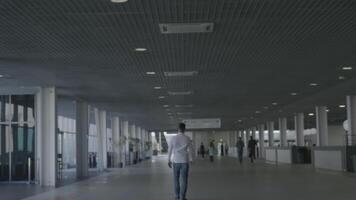 The businessman walks in the business center through the long corridor. HDR. Rear view of a young adult man in white shirt and jeans walking inside white building interior. video