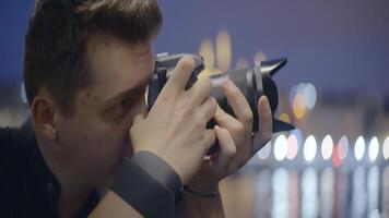 Young man takes pictures with camera on background of night city. Action. Handsome young man with professional camera on blurry city background in evening video