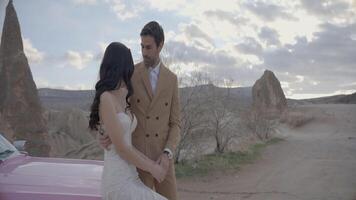 hermosa Pareja con coche en antecedentes de montañas. acción. elegante trajes de recién casados de vacaciones en montañas. elegante recién casados sentar en capucha de coche en antecedentes de cañón video