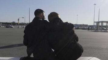 giovane gay maschio coppia seduta su un' panchina a autobus fermare, parlando, e abbracciare. hdr. Due uomini nel amore in attesa per un' autobus all'aperto durante conversazione su un' soleggiato giorno, concetto di lgbt relazioni. video