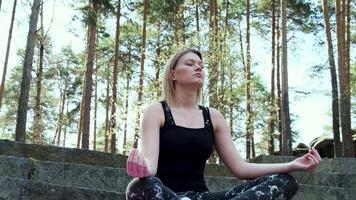 Happy woman deeply connected with nature feels unity and meditating in forest outdoors. Concept. Blond girl in sports suit sitting on concrete stairs in meditation on a summer day. video