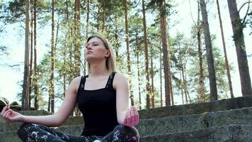 contento mujer profundamente conectado con naturaleza siente unidad y meditando en bosque al aire libre. concepto. rubio niña en Deportes traje sentado en hormigón escalera en meditación en un verano día. video