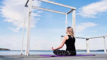 blond ung kvinna Sammanträde i lotus placera nära sjö och håller på med andas övningar. begrepp. flicka i sporter kostym Sammanträde i meditation på en pir förbi de vatten på blå molnig himmel bakgrund. video