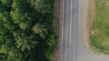 View from a helicopter . Scene . A narrow road on which a cargo truck with a blue front and a car with a trailer for transportation are traveling , there is a forest and a road with grass nearby . video
