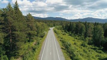 Top view of truck driving along highway in forest area. Scene. Truck is driving along highway among green forest on background of mountains. Truck goes along route in forest area on sunny summer day video