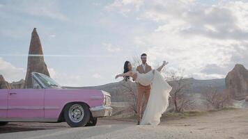 glücklich Hochzeit Paar im Berge posieren im Vorderseite von ein Kamera. Aktion. Bräutigam im passen halten schön Braut im seine Waffen in der Nähe von das Cabriolet Rosa Wagen. video