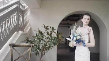 donna in posa con fiori. azione. riflessivo giovane sposa nel bellissima lungo nozze vestito Tenere mazzo e in piedi vicino il Casa con scale. video