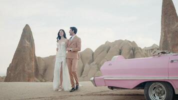 Beautiful young couple during photo session on the top of a mountain in Cappadocia. Action. Romantic love of man and woman couple near the cabriolet pink car. video