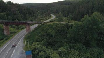 An abandoned rusty railroad bridge above the curving road with a driving truck. Scene. Green summer forest and asphalt bended road under the bridge with supporting columns. video