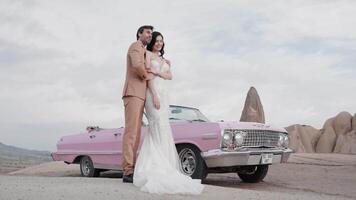 Elegant wedding photo shoot. Action. A beautiful bride in a wedding dress with a groom in a brown suit posing for a photographer on camera with happy faces in the desert next to a pink small car video