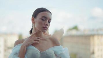 Young beautiful bride in a white wedding dress standing on a balcony on blurred city background. Action. Elegant woman with the waving silk scar tied around her ponytail. video