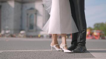 Legs of stylish bride and groom on the background of city summer street with driving cars. Action. Man in black suit and a woman in white dress. video