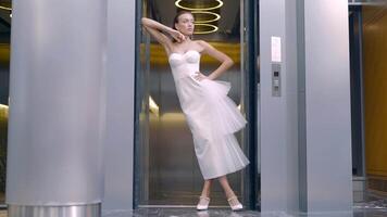 Woman in white dress poses on background of lift. Action. Elegant young woman poses inside modern building. Beautiful woman in elegant white dress posing video