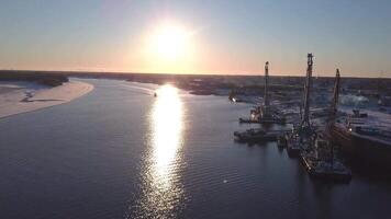 Moored cargo transport ships and barges with crains on a wide river at summer dawn. Clip. Aerial view of water transport against shining sun. video