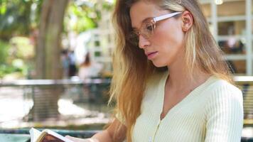 Woman in eyeglasses reading book sitting at table in outdoor cafe video