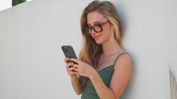 sorridente donna nel di moda occhiali utilizzando smartphone pendente su parete video