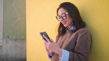 leende kvinna använder sig av smartphone mot gul vägg utanför video