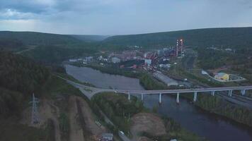 un aéreo zumbido paisaje de un pequeño pueblo con río y verde boscoso montañas. acortar. un puente cruce dos río bancos en nublado cielo antecedentes. video