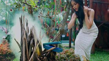 mujer en blanco vestir astillas un fuego al aire libre con fumar creciente alrededor, evocando un rústico y natural ambiente video