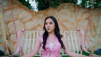 Smiling young woman in a pink dress sitting on a swing with a rustic wall in the background. video