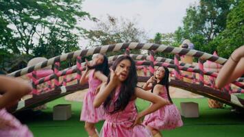 Three girls in pink dresses running hand in hand in a park with a whimsical oversized bridge in the background. video