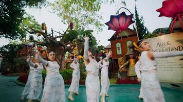 Group of women in white dresses performing a dance at an outdoor event with large floral decorations in the background. video