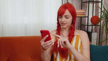 Cheerful ginger woman sitting on sofa, using smartphone share messages on social media application video