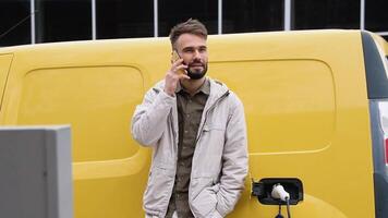 lächelnd Mann im beiläufig tragen reden Telefon während gelehnt auf seine Ladung elektrisch Auto, Stehen auf das Laden Bahnhof zum Laden ein Wagen. elektrisch Auto Laden Konzept video