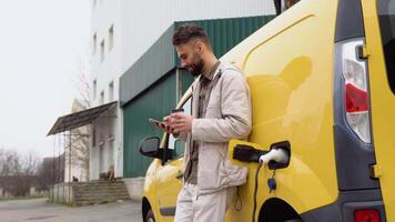 jovem turco homem conversando em Móvel telefone e bebidas café enquanto elétrico carro é cobrando em ao ar livre estação video