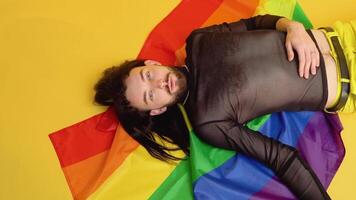 Gay man dressed in mesh t-shirt lies on yellow background with a rainbow flag. Concept diversity, transsexual, and freedom video