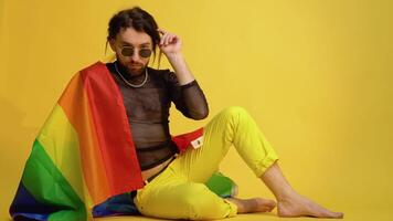 Gay man dressed in mesh t-shirt and sunglasses sits on yellow background with a multicolored rainbow flag. Concept diversity, transsexual, and freedom video