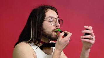 Young sexy gay man paints lips with green lipstick isolated on red background. People lifestyle fashion lgbtq concept video