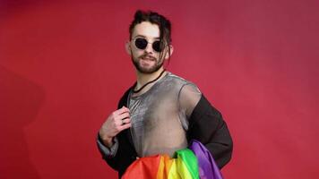Transsexual man dressed in mesh t-shirt and sunglasses on red background with a multicolored rainbow flag. Concept diversity, transsexual, and freedom video