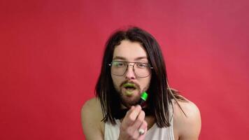 Young sexy gay man paints lips with green lipstick isolated on red background. People lifestyle fashion lgbtq concept video
